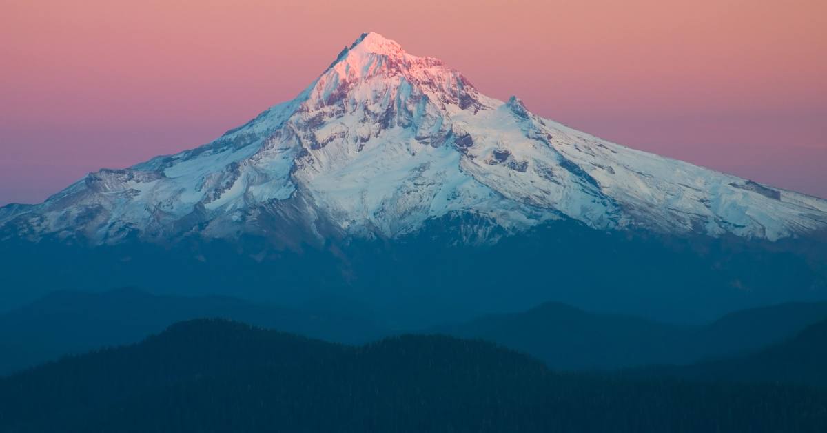 Mount Hood at sunset, its peak lit pink above dark blue forested ridges